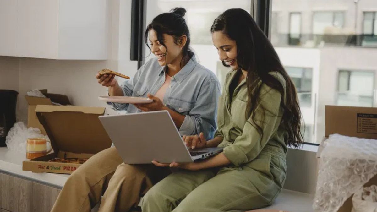 Two happy friends sitting next to each other, one eating pizza and the other using a laptop.